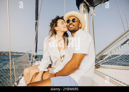 Una coppia gioiosa che si bacierà su una barca a vela, godendosi una fuga romantica sull'oceano. Mostrano felicità e relax sotto il cielo soleggiato, epitomizin Foto Stock