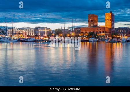 Splendida vista serale del municipio di Oslo e di Aker Brygge dal molo di Akershus, con edifici illuminati che si riflettono sulle acque calme sotto una suggestiva S. Foto Stock