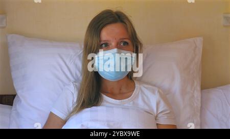 Giovane donna in maschera medica che riposa a letto, mostrando precauzione durante Foto Stock