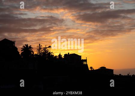 Silhouette di case e palme su una collina al tramonto a Puerto de la Cruz, Tenerife, con un cielo arancione e giallo vibrante. Foto Stock