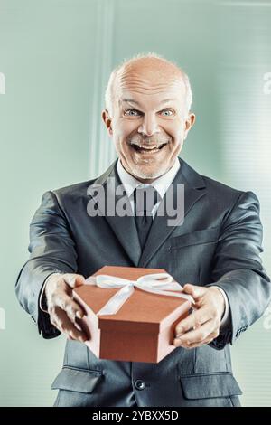 Un uomo d'affari maturo che indossa un abito e una cravatta regala un regalo con un grande sorriso, desideroso di regalarlo a qualcuno di speciale Foto Stock