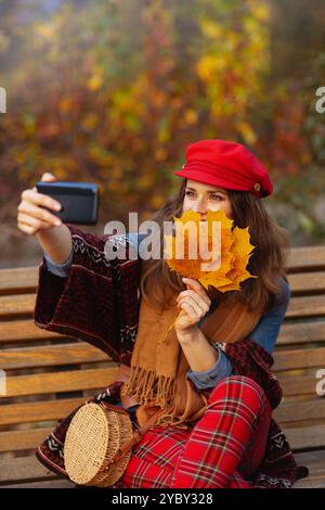 Ciao autunno. elegante ragazza sorridente in cappello rosso con foglie autunnali, sciarpa e borsa per incontri virtuali seduti in panchina nel parco cittadino. Foto Stock