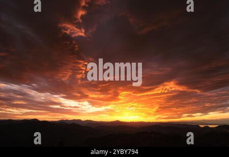 paesaggio di un tramonto in cui si può vedere il sole che si nasconde tra le montagne sullo sfondo mentre più vicino le nuvole nel cielo si illuminano di arancione a. Foto Stock