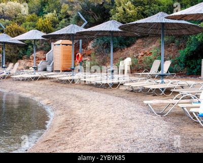 File di lettini vuoti si trovano sotto ombrelloni di paglia su una spiaggia deserta, invitando al relax in riva al mare tranquillo Foto Stock