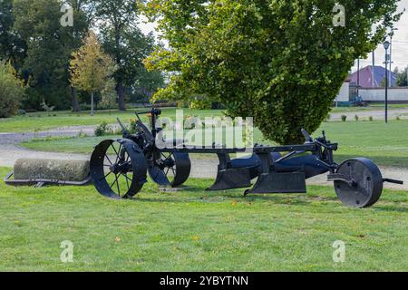 Aratro agricolo d'epoca con grandi ruote esposte su un campo erboso in un parco circondato da alberi in una giornata di sole Foto Stock