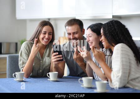 Quattro felici amici interrazziali che incrociano le dita controllando il telefono in cucina a casa Foto Stock