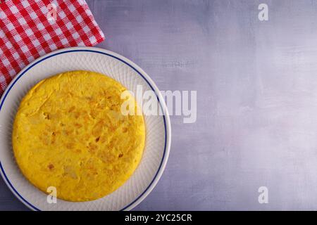 Goditi una deliziosa frittata di patate spagnole accanto a un affascinante tovagliolo a scacchi rosso, adagiato su un fresco tavolo blu in cemento in questa invitante scena Foto Stock