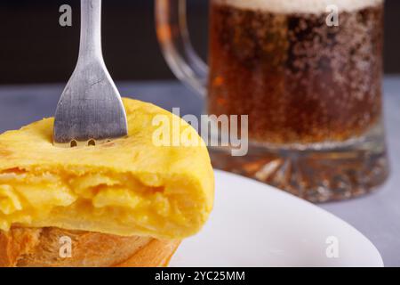 Spiedino di omelette spagnole trafilate con una forchetta, su una fetta di pane, accanto a una tazza di birra sullo sfondo, appoggiata su un tavolo di cemento blu Foto Stock