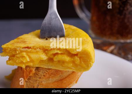 Primo piano dello spiedino di tortilla spagnola con fetta di pane, forchetta che pierta la frittata, con una tazza di birra fredda sullo sfondo, su un tavolo di cemento blu Foto Stock