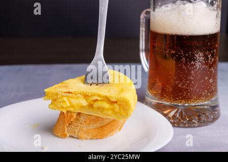 Spiedino parziale di omelette spagnole con pane e forchetta, con una tazza di birra sullo sfondo, posto su un tavolo di cemento blu, in una vista dettagliata Foto Stock