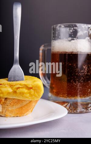 Forchetta che strizza una fetta di tortilla spagnola sul pane, con una birra fredda sullo sfondo, su una superficie di cemento bluastro, scattata in dettaglio Foto Stock