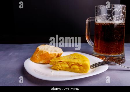 Vista dettagliata dello spiedino di tortilla spagnolo con fetta di pane, forchetta che lo perfora, con una tazza di birra fredda sullo sfondo, su un tavolo di cemento bluastro Foto Stock