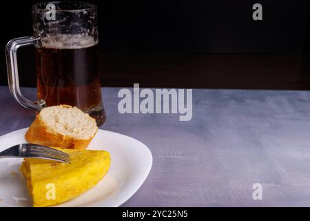 Spiedino di omelette spagnole su una fetta di pane, tenuto da una forchetta, con una tazza di birra delicatamente messa a fuoco sullo sfondo, su una superficie blu di cemento Foto Stock