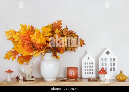 Foglie autunnali gialle e arancioni brillanti in vaso bianco su piano in legno. Case decorative nelle vicinanze e candele su sfondo bianco in gesso. Autunno d Foto Stock