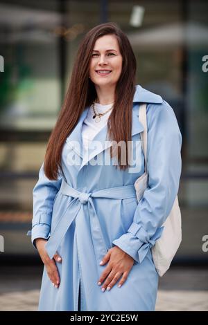 Ritratti donna di taglia più affascinante con i capelli lunghi sorride con sicurezza all'aperto, indossa il trench azzurro alla moda e porta borsa di tela e showcas Foto Stock