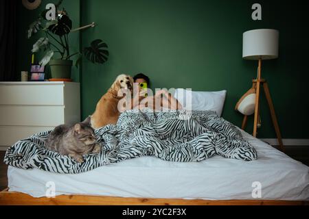 Un giovane giace a letto con un cane e un gatto, il telefono tra le mani Foto Stock