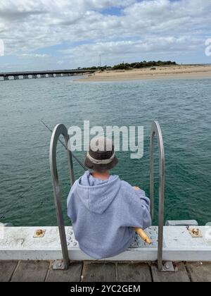 Ragazzo seduto sul bordo del molo con una canna da pesca Foto Stock