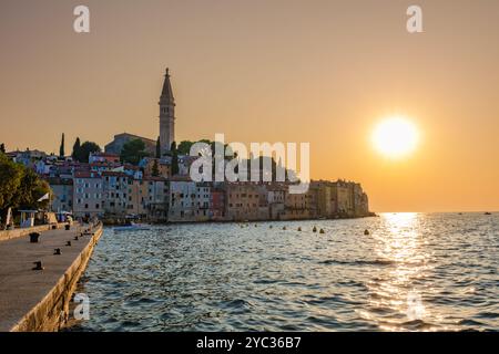 Mentre il sole tramonta su Rovigno, le vivaci sfumature danzano sull'acqua calma, illuminando gli edifici storici lungo la costa. L'iconico campanile Foto Stock