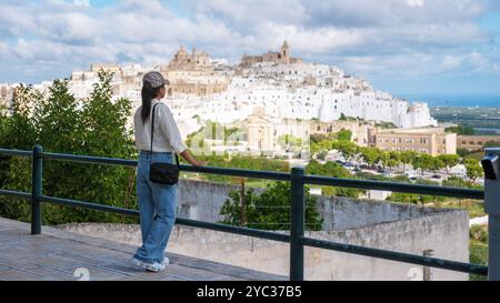 Un viaggiatore si trova in un punto panoramico, ammirando gli edifici imbiancati di Ostuni, in Italia, annidati su una collina sotto un cielo nuvoloso. Il verde sottostante esalta la pittoresca bellezza della città. Foto Stock