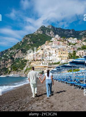 Un paio di cammina mano nella mano lungo la pittoresca spiaggia di Positano, circondata da case colorate sulla scogliera e dal tranquillo Mar Mediterraneo. Il cielo è luminoso e l'atmosfera è tranquilla. Foto Stock
