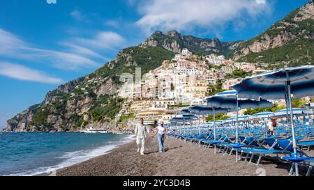 Due persone passeggiano mano nella mano lungo la spiaggia di ciottoli della costiera amalfitana, con le vivaci case sulla scogliera di Positano che si innalzano maestosamente sopra di loro sotto un cielo azzurro. Foto Stock