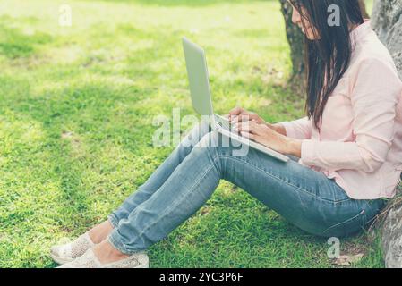 Donna asiatica seduta parco verde con computer portatile. Donna che lavora in un'azienda felice di un notebook che utilizza un notebook con le mani che digitano sulla tastiera Foto Stock