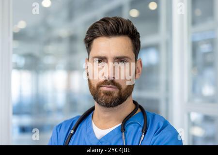 Medico maschile sicuro di sé con barba in scrub blu e stetoscopio in ambiente ospedaliero moderno. Il contatto diretto con gli occhi trasmette professionalità, dedizione, competenza e fiducia nel campo medico. Foto Stock