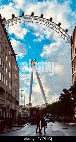 Una vista panoramica del London Eye incorniciata tra gli edifici della città con un cielo nuvoloso, che mostra la vita urbana e una strada trafficata con pedoni e veicoli Foto Stock