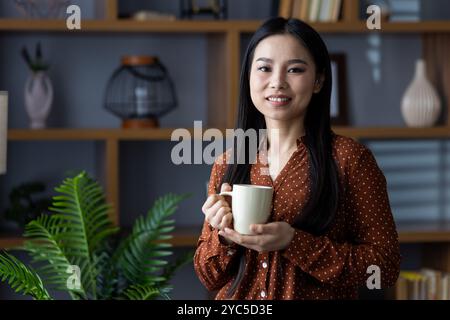 Donna asiatica sorridente che tiene una tazza di caffè in un ambiente accogliente. Rilassato e allegro con camicia a pois marrone, godendosi del tempo libero. Caldo e invitante Foto Stock