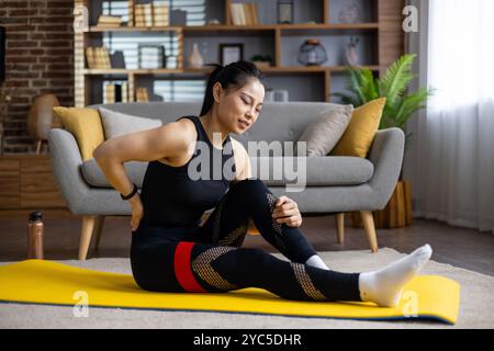 Donna asiatica che soffre mal di schiena seduta su un tappetino da yoga in salotto. Indossando abiti da allenamento neri, si allunga delicatamente per il sollievo. Concentratevi sulla salute AN Foto Stock