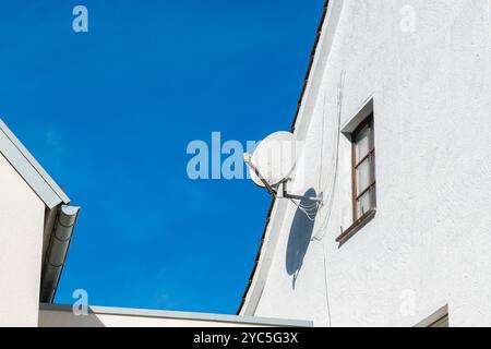 Una parabola satellitare è installata sul lato di un edificio bianco, in netto contrasto con il cielo azzurro. La configurazione è tipica per le comunicazioni moderne Foto Stock