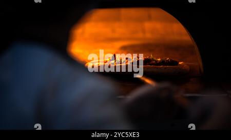 Una tradizionale pizzeria italiana, uno chef in uniforme posiziona con cura una pizza con una crosta marrone dorata su una pietra in un forno a legna. Le fiamme bla Foto Stock