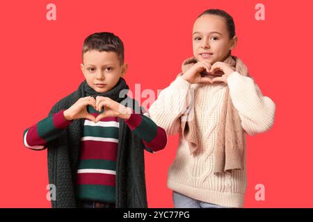 Bambini piccoli e felici con abiti invernali caldi che mostrano gesti del cuore su sfondo rosso Foto Stock