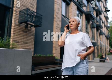 Ritratto ad angolo basso di una donna caucasica anziana che parla su smartphone in posizione su sfondo urbano con illuminazione soffusa. Foto Stock