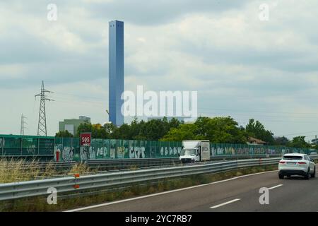 Brescia, Italia - 13 giugno 2023: I viaggiatori navigano su un'autostrada trafficata delimitata da recinzioni verdi adornate da colorati graffiti, mentre un suggestivo towe blu Foto Stock