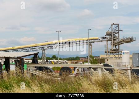 Brescia, Italia - 13 giugno 2023: I macchinari alti incombe sul paesaggio, con nastri trasportatori che trasportano i materiali mentre i serbatoi di stoccaggio e il verde p Foto Stock