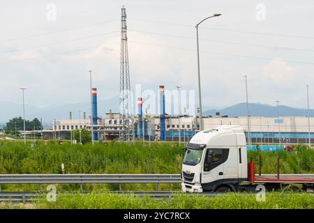 Brescia, Italia - 13 giugno 2023: Un camion commerciale bianco naviga sull'autostrada, passando davanti a un vasto sito industriale adornato da macchie colorate Foto Stock