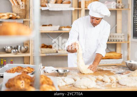 il panettiere maschile si trova sul banco di lavoro, impastando e modellando l'impasto per preparare pane, croissant e baguette Foto Stock