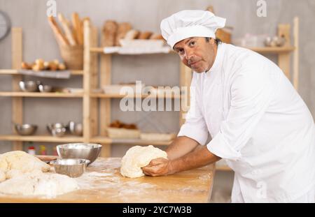 il panettiere maschile si trova sul banco di lavoro, impastando e modellando l'impasto per preparare pane, croissant e baguette Foto Stock