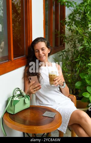 Ritratto di una donna russa di 39 anni senza trucco seduta con un bicchiere di caffè ghiacciato in abito bianco Foto Stock