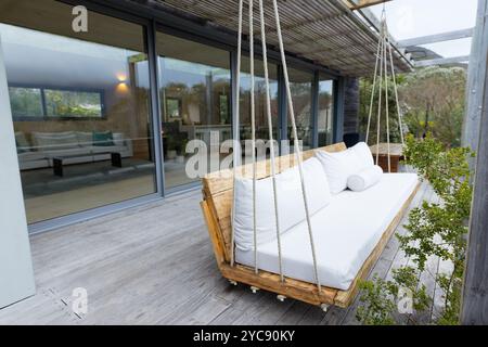 Divano a dondolo in legno sul moderno patio con porte in vetro e vegetazione lussureggiante, a casa Foto Stock