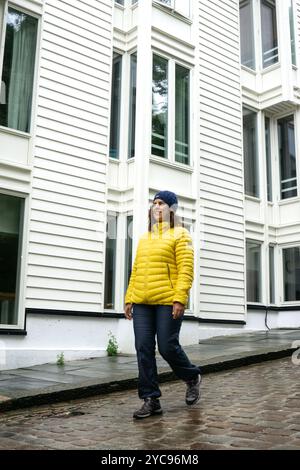 Una giovane donna bruna, di circa 30 anni, cammina di fronte a un edificio bianco in una strada di ciottoli bagnata con il bel tempo, indossando un anorak giallo Foto Stock