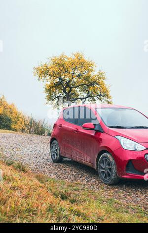 Auto rossa su strada ghiaiosa in montagna in giornata nebbiosa. Viaggio autunnale in auto. Auto Hatchback nel fango in montagna con nuvole. Viaggio rurale in auto. Foto Stock