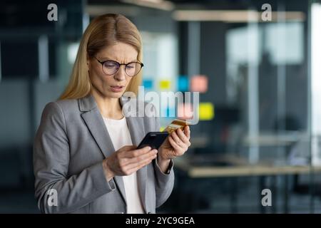 Donna d'affari con gli occhiali in ufficio, con carta di credito e telefono, che sembra preoccupata. Utilizzo di tecnologie moderne, possibile problema di pagamento online o identità Foto Stock