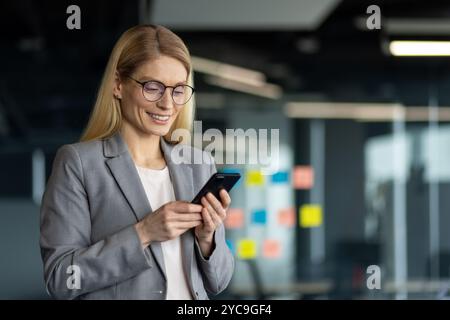 Donna d'affari matura in ufficio che controlla il telefono. Sta indossando occhiali e un vestito grigio. Sorridendo con fiducia, trasuda successo, professionalità e. Foto Stock