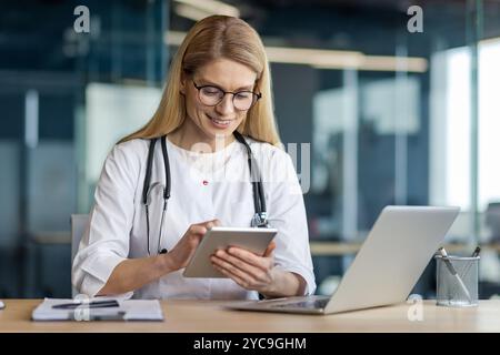 Medico sorridente che usa un tablet in ufficio. Indossare cappotto bianco, stetoscopio, occhiali, lavorare su un computer portatile. Rappresenta il settore sanitario, tecnologico, professionale Foto Stock