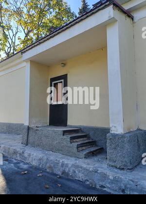 Una piccola entrata con scalini di cemento che conduce ad una porta di legno scuro, incorniciata da una semplice facciata gialla. La scena riflette l'invecchiamento dell'infrastruttura in difficoltà Foto Stock