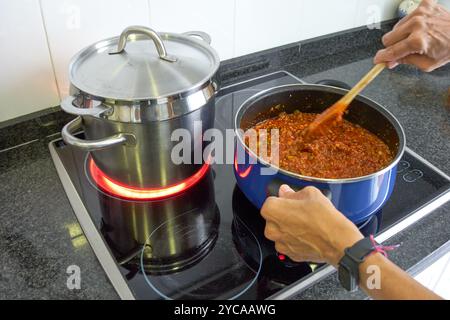 Due pentole per preparare i maccheroni bolognesi in una stufa elettrica domestica. Una pentola con la salsa di pomodoro e un'altra con la cottura della pasta con una luce brillante Foto Stock