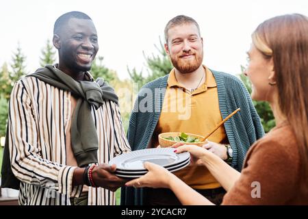 In vita ritratto di un gruppo di amici che prepara un tavolo da pranzo durante la festa insieme e che serve piatti incentrati sull'uomo afroamericano sorridente che tiene i piatti in mano Foto Stock