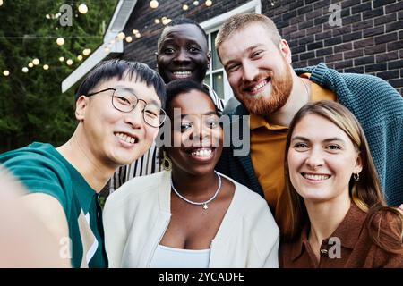 POV di un gruppo multietnico di amici che sorride felicemente alla macchina fotografica scattando selfie insieme all'aperto nel cortile della casa Foto Stock